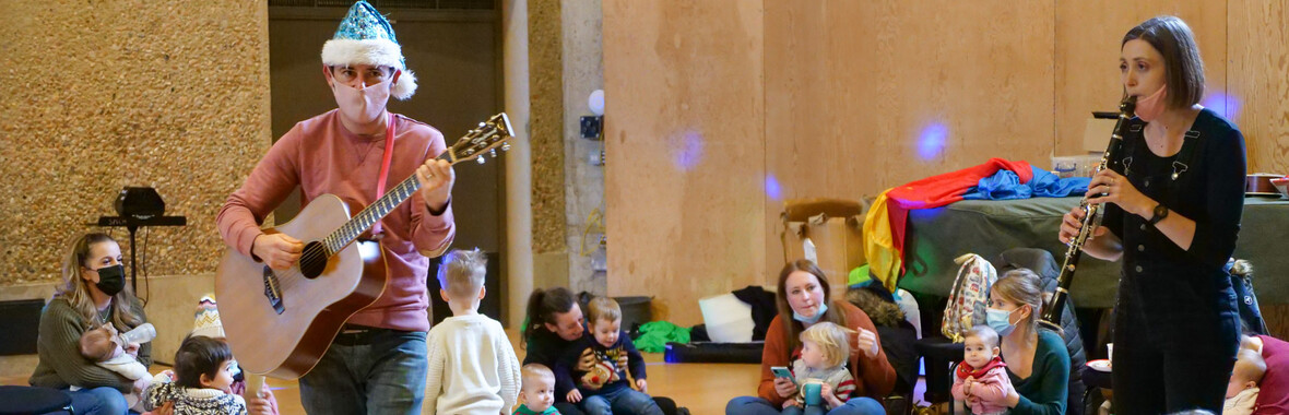 A man in a green santa hat playing guitar to an audience of children with their parents.