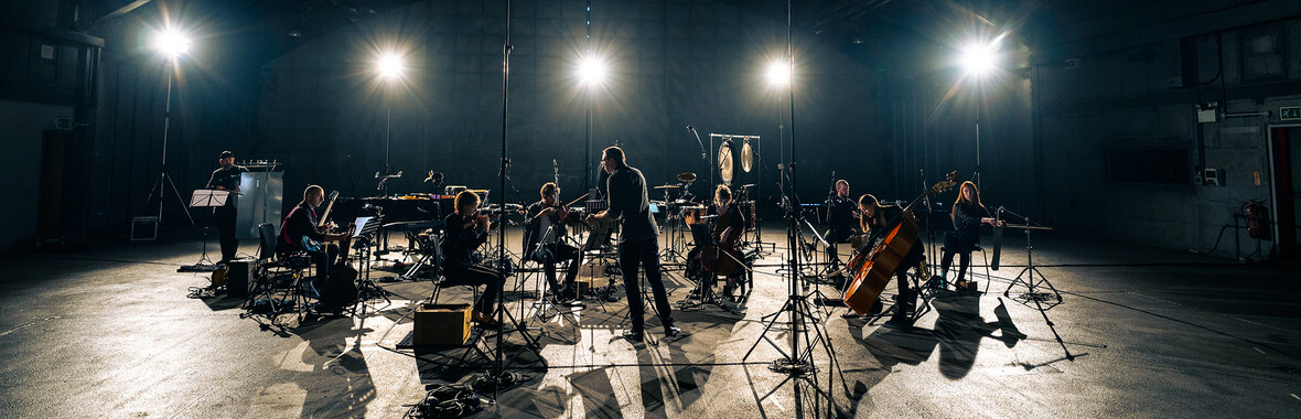 The musicians of Riot Ensemble playing in a dark space with flares from bright stage lights